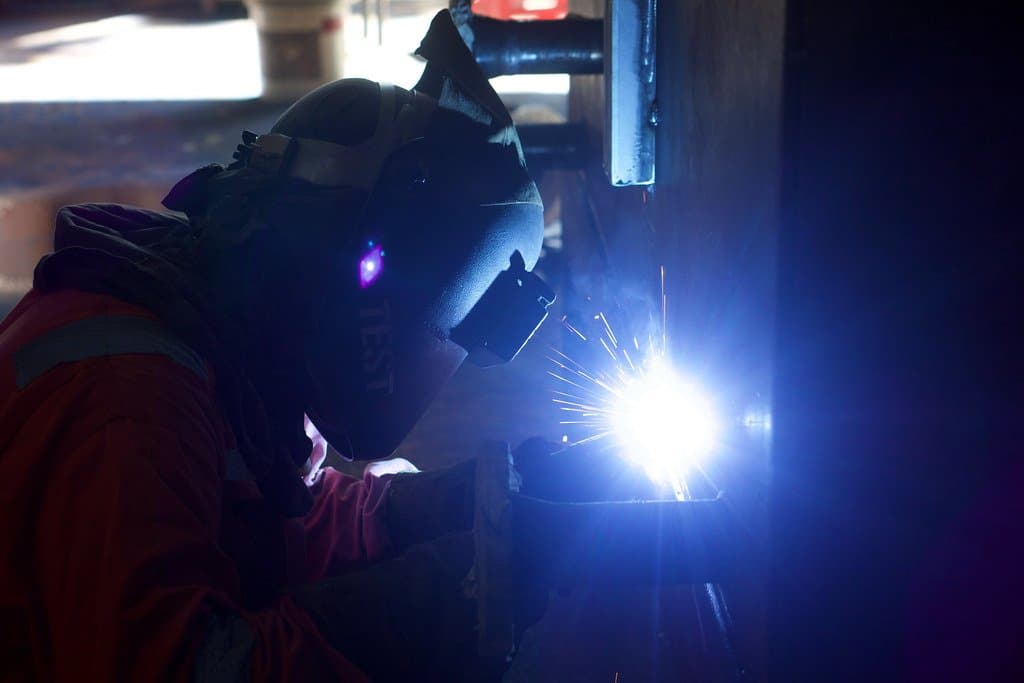 A welder in protective gear working on metal, with visible sparks, in an industrial facility